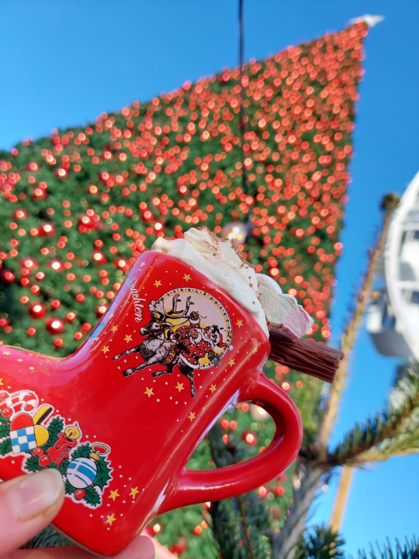 Festive red boot mug of hot chocolate topped with cream in front of the lit Christmas tree