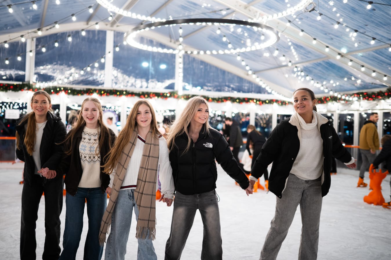 Five friends skating hand in hand under festive lights at Oaks on Ice